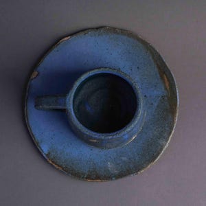 May include: A blue ceramic cup and saucer set with a rustic, hand-thrown look. The cup has a rounded shape and a small handle.