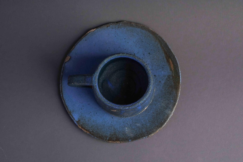 May include: A blue ceramic cup and saucer set with a rustic, hand-thrown look. The cup has a rounded shape and a small handle.