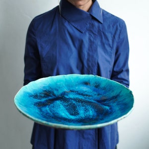 Pode incluir: Uma grande travessa redonda de cerâmica com um vibrante esmalte turquesa e azul escuro. A borda tem uma borda ligeiramente irregular, e as cores se misturam para criar um design artístico único. A travessa é segurada contra uma camisa azul.