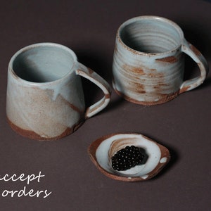 May include: Two ceramic mugs with a light blue glaze and brown accents. The mugs have a rustic, handmade look. A small ceramic dish with a light blue glaze and brown accents holds a cluster of black berries.