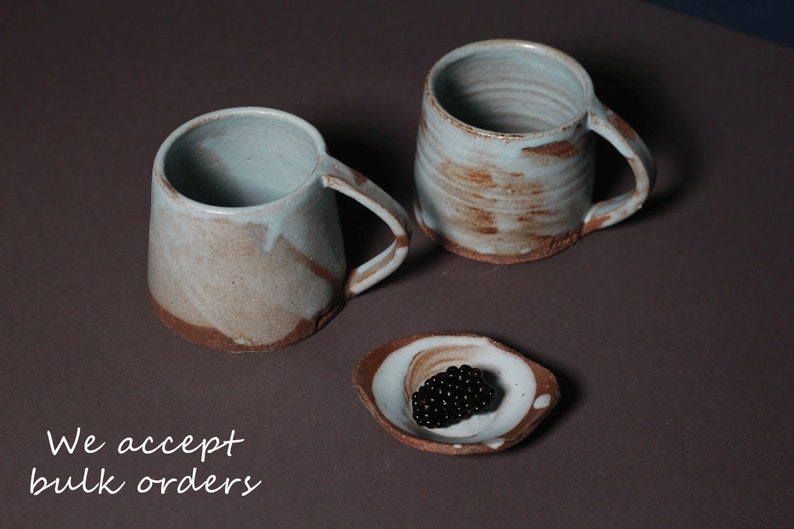 May include: Two ceramic mugs with a light blue glaze and brown accents. The mugs have a rustic, handmade look. A small ceramic dish with a light blue glaze and brown accents holds a cluster of black berries.