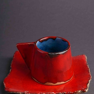 May include: A red ceramic coffee cup with a blue glaze inside and a matching red saucer. The cup has a unique, angular handle.