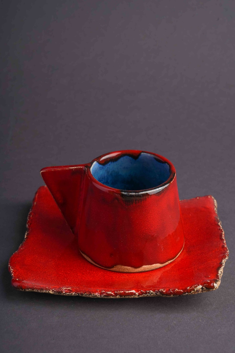 May include: A red ceramic coffee cup with a blue glaze inside and a matching red saucer. The cup has a unique, angular handle.