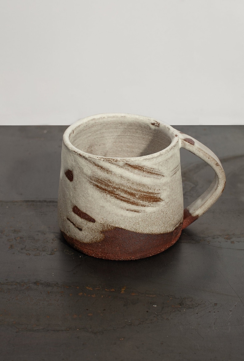 May include: Hand-thrown ceramic coffee mug with matte cream upper glaze and rich, earthy brown base. Features organic brushstroke patterns, a gently tapered profile, and a sturdy curved handle. Its artisanal texture and warm tones evoke a cozy, rustic farmhouse aesthetic.