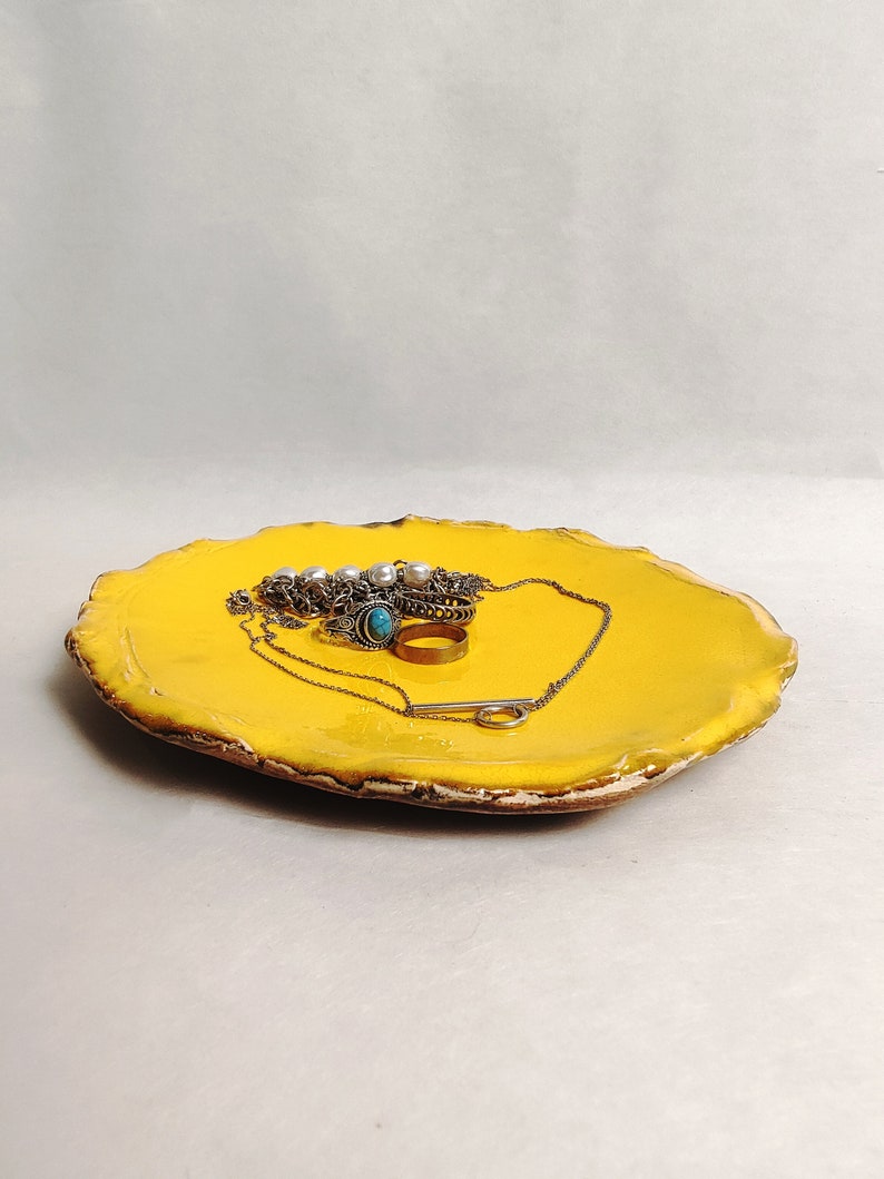 Puede incluir: Un plato de cer&aacute;mica amarillo con un borde ondulado. El plato est&aacute; lleno de joyas, incluyendo un collar, un anillo y una pulsera.