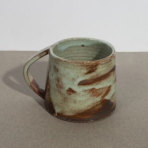 May include: A handmade ceramic mug with a light green glaze and brown speckles. The mug has a rounded shape and a handle.
