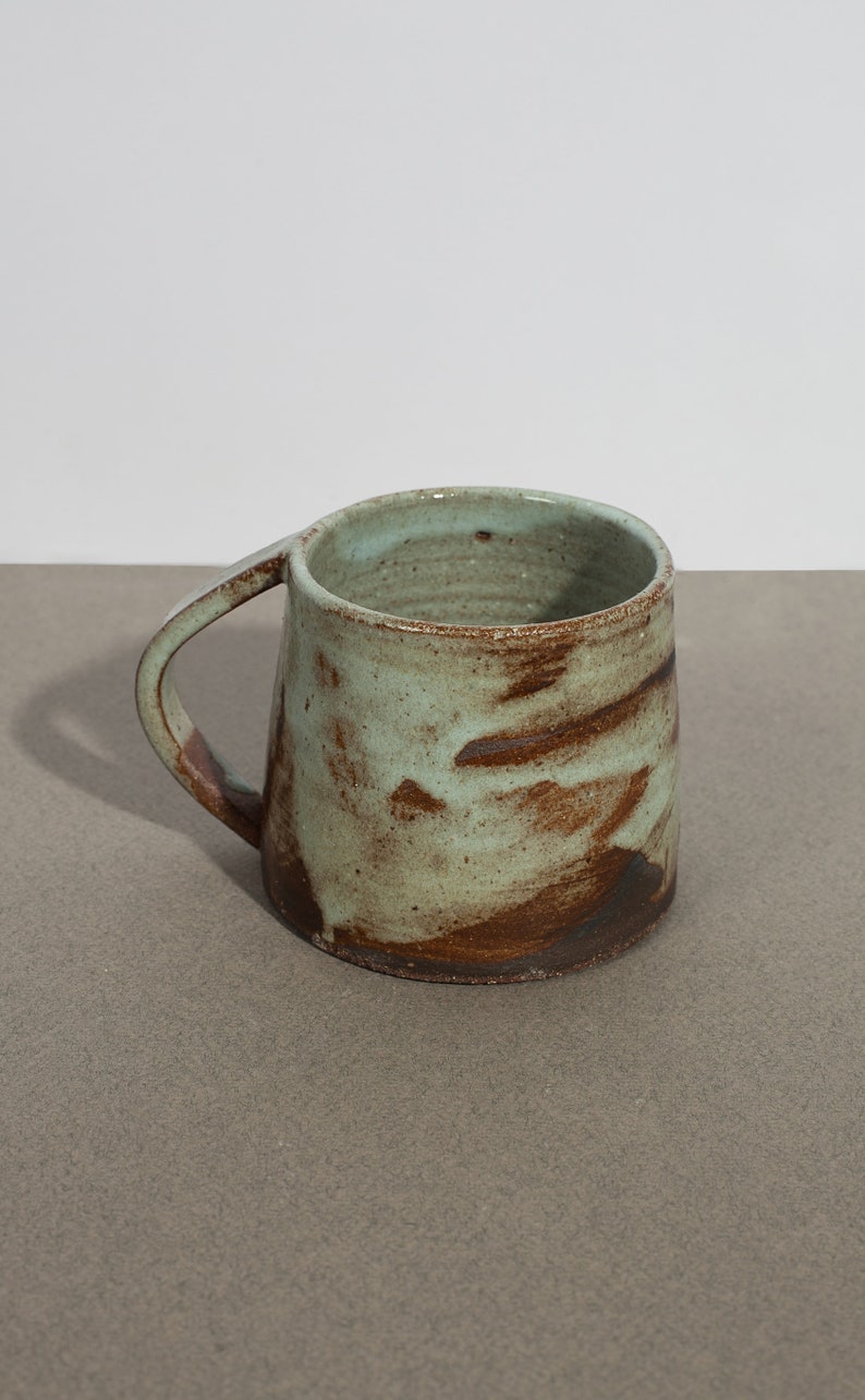 May include: A handmade ceramic mug with a light green glaze and brown speckles. The mug has a rounded shape and a handle.
