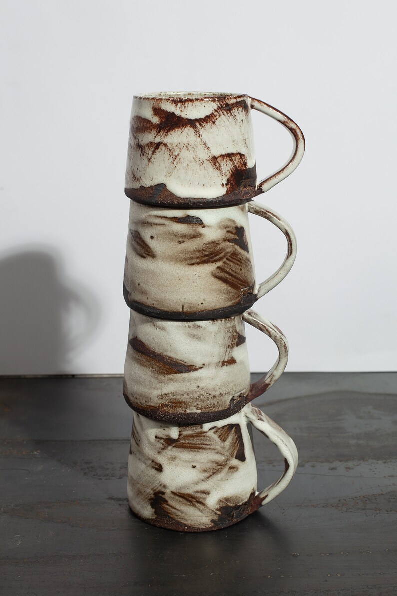 May include: A stack of four ceramic mugs with a white glaze and brown brushstrokes. The mugs have a rounded shape and a handle on the side.