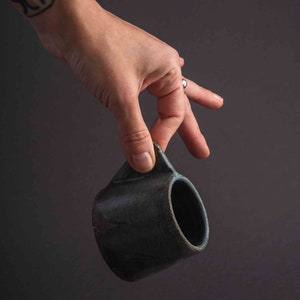 May include: A hand holds a dark blue ceramic mug with a rounded shape and a slightly flared lip.