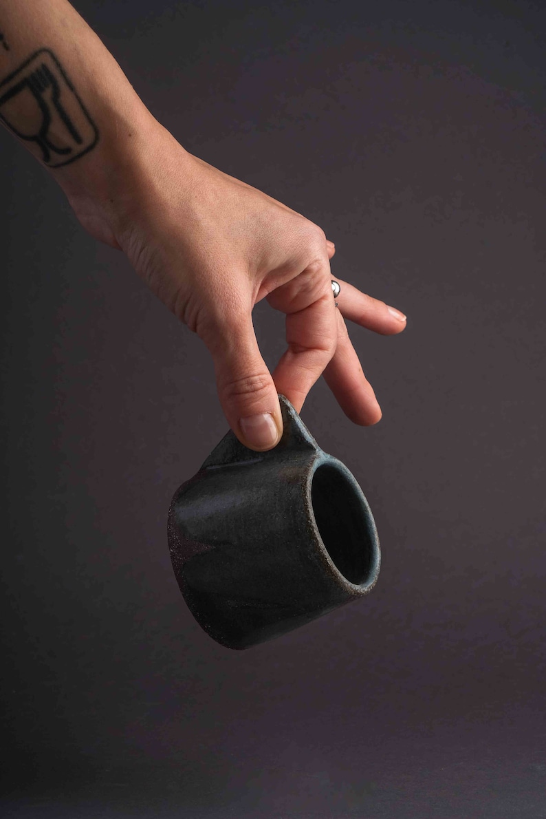 May include: A hand holds a dark blue ceramic mug with a rounded shape and a slightly flared lip.