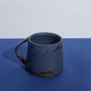 May include: A blue ceramic mug with a speckled glaze and a brown rim. The mug has a unique, slightly asymmetrical shape and a handle.