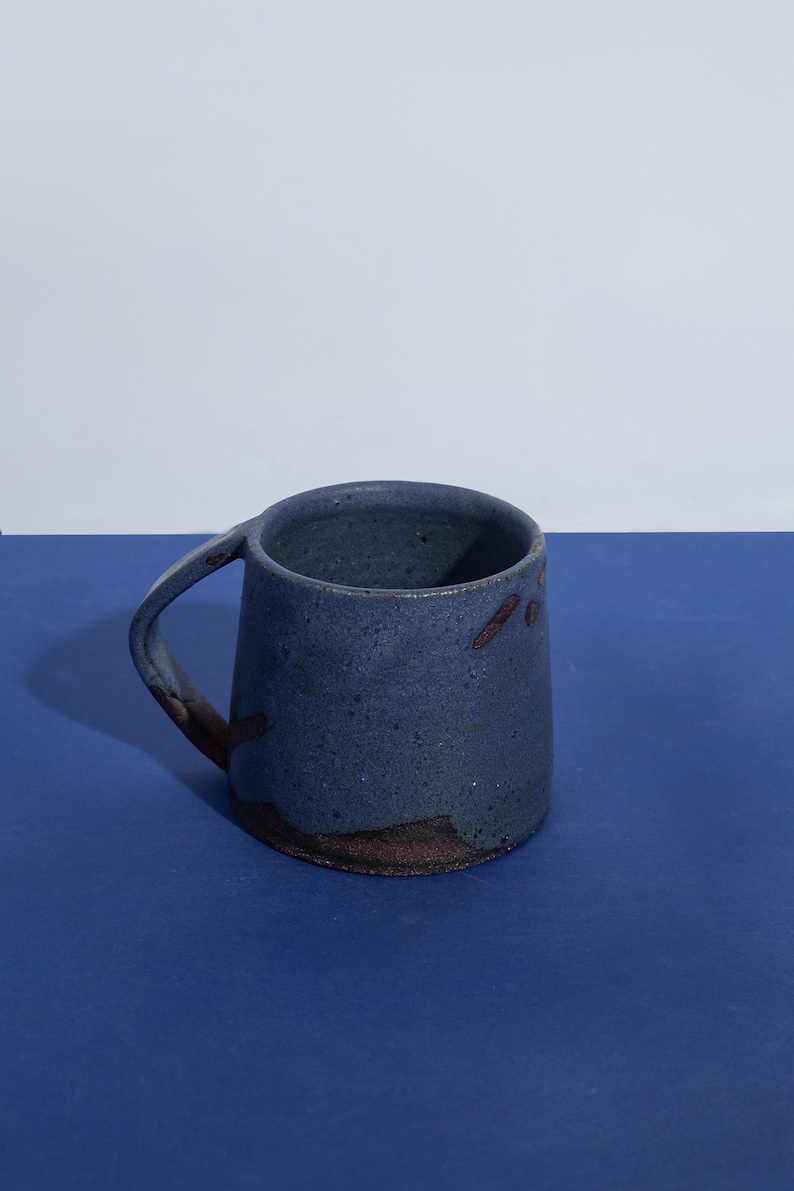 May include: A blue ceramic mug with a speckled glaze and a brown rim. The mug has a unique, slightly asymmetrical shape and a handle.