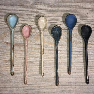 May include: Seven ceramic spoons in various colors, including blue, pink, white, and black. The spoons are all hand-painted and have a rustic, handmade look.