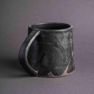 May include: A black ceramic mug with a textured, uneven surface and a wide, rounded handle. The mug has a dark grey background with subtle hints of brown and purple.