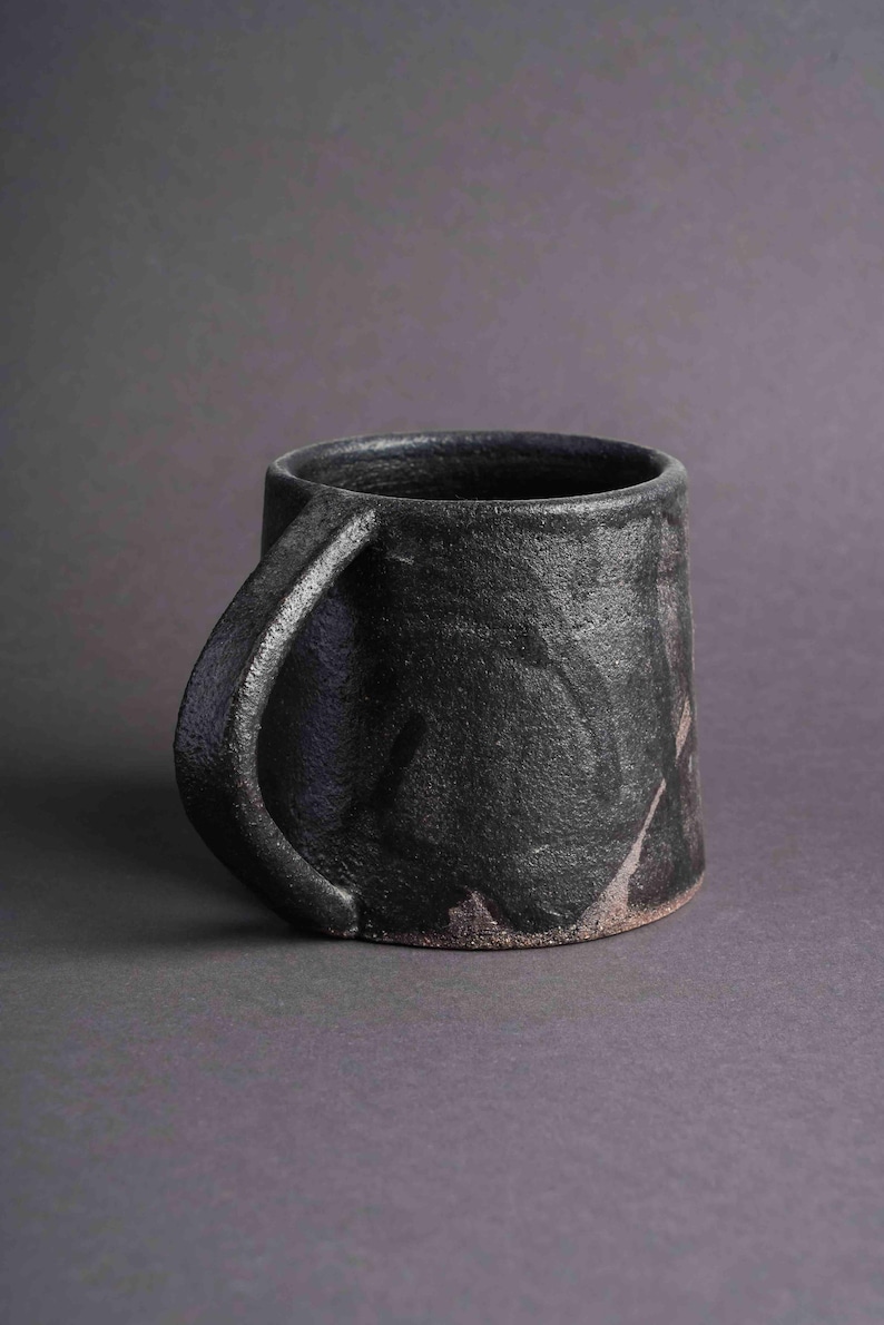 May include: A black ceramic mug with a textured, uneven surface and a wide, rounded handle. The mug has a dark grey background with subtle hints of brown and purple.