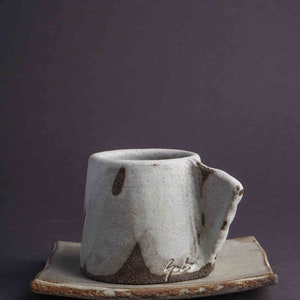 May include: A white ceramic coffee cup with a brown glaze and a unique, asymmetrical shape. The cup is sitting on a matching saucer.
