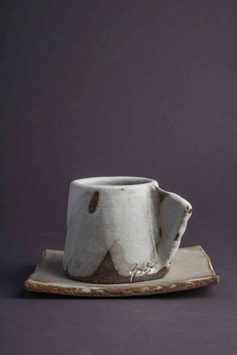 May include: A white ceramic coffee cup with a brown glaze and a unique, asymmetrical shape. The cup is sitting on a matching saucer.