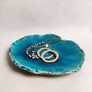 Puede incluir: Plato de cer&aacute;mica azul verdoso con un borde rugoso y texturizado. El plato tiene un borde dorado y est&aacute; decorado con un dise&ntilde;o de espiral dorado. Dos collares de oro descansan sobre el plato.