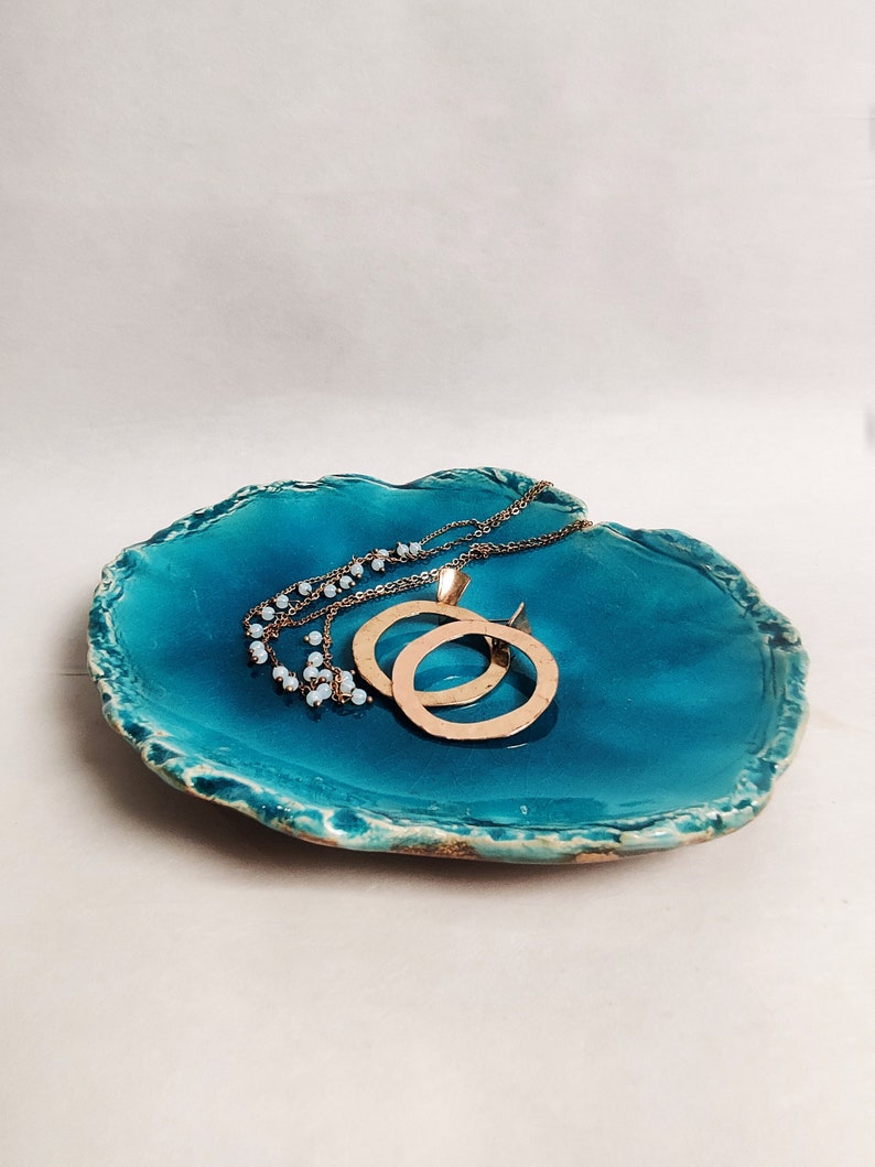 Puede incluir: Plato de cer&aacute;mica azul verdoso con un borde rugoso y texturizado. El plato tiene un borde dorado y est&aacute; decorado con un dise&ntilde;o de espiral dorado. Dos collares de oro descansan sobre el plato.