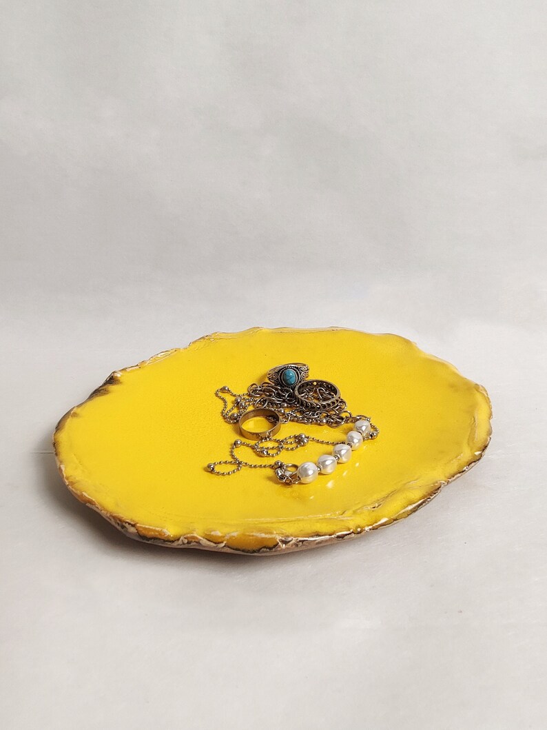 Puede incluir: Un plato de cer&aacute;mica amarillo para joyas con un borde &aacute;spero e irregular. El plato est&aacute; lleno de joyas, incluyendo un collar de cadena con detalles de perlas y varios anillos.