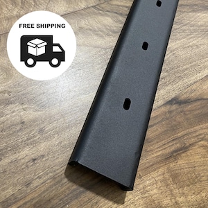 Könnte beinhalten: Ein langer, schwarzer Metallstab mit gleichmäßig verteilten Löchern. Der Stab ist abgewinkelt und zeigt seine rechteckige Form und matte Oberfläche. Der Hintergrund ist eine Holzoberfläche.