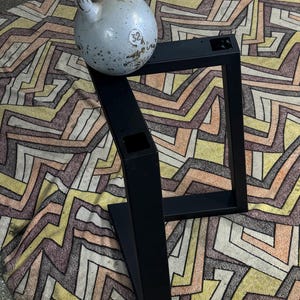 Könnte beinhalten: Schwarze Metalltischbeine mit quadratischem Design. Die Beine stehen auf einem gemusterten Teppich mit einer Kettlebell auf den Beinen.