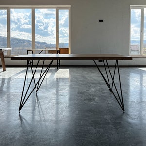 Könnte beinhalten: Ein rechteckiger Holztisch mit einer dunkelbraunen Oberfläche und schwarzen Metall-Haarnadelbeinen. Der Tisch steht in einem hellen Raum mit großen Fenstern mit Blick auf eine Landschaft. Der Tisch ist etwa 183 cm lang.