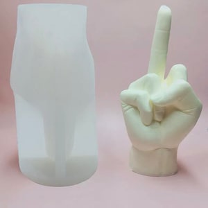 May include: A white silicone mould of a hand giving the middle finger. The mould is on a pink surface.