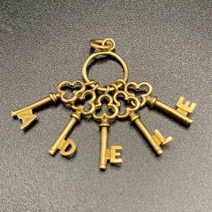 Puede incluir: Un amuleto dorado con un grupo de seis llaves, cada una con una letra en el extremo. Las letras deletrean "ADORE" y el amuleto tiene un anillo en la parte superior para sujetarlo a un collar o pulsera.
