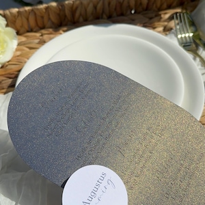 May include: A gray, oval-shaped menu card with a glittery finish. The menu card features a handwritten menu with the text "Pruttle Arancini, Parmesan, Seared Scallops, Lemon Butter, Crab Tartlet, Chive Creme" and "Salude, Heirloom Tomato, Burrata, Basil, Black Olive, Champagne Vinaigrette, Truffle, Parmesan, Anchovy, Gem, Parsley". A white, round card with the text "Augustus Hemming" is placed on top of the menu card.