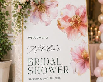 Puede incluir: Un letrero rectangular blanco con marco dorado sobre un caballete dorado, con flores rosas estilo acuarela y el texto: WELCOME TO Natalia's BRIDAL SHOWER SATURDAY, JULY 20, 2035. Un arreglo floral de rosas rosas y follaje se encuentra arriba.