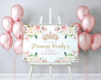 May include: A sign on an easel reads "Welcome to Princess Wendy's First Birthday" in gold script, surrounded by floral arrangements and a gold crown. Pink balloons are clustered on either side of the sign, creating a festive atmosphere.