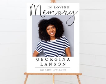 May include: A white memorial sign with a black border and the text "In Loving Memory" in a cursive font. A photo of a woman with curly black hair is in the center of the sign. The text "Georgina Lanson" is below the photo, and the dates "July 1, 2000 - April 3, 2090" are below the name.