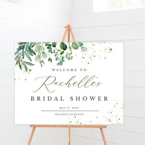 May include: A white welcome sign with green foliage and gold glitter accents. The sign reads "Welcome to Rachelle's Bridal Shower May 17, 2025 #RachelleInLove"