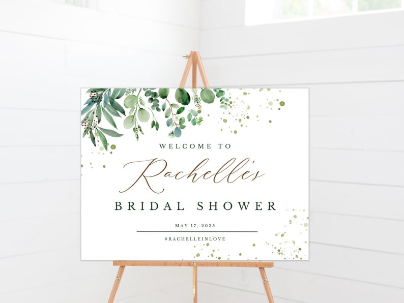 May include: A white welcome sign with green foliage and gold glitter accents. The sign reads "Welcome to Rachelle's Bridal Shower May 17, 2025 #RachelleInLove"
