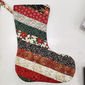 May include: A red, green and black quilted Christmas stocking with a diagonal striped pattern. The stocking has a loop at the top for hanging.