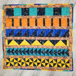 May include: A colourful quilt with a geometric pattern. The quilt features rows of teal squares, orange rectangles, blue triangles, yellow triangles, and teal triangles. The quilt is bordered with a brown and yellow fabric.