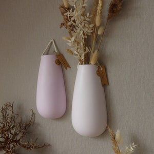 Könnte beinhalten: Zwei Keramik-Wandvasen in hellrosa und weiß. Die Vasen sind tropfenförmig und haben oben einen Seilgriff. Die Vasen hängen an einer Wand und sind mit Trockenblumen gefüllt.