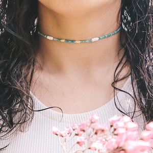 Peut inclure: Gros plan d'une femme portant un collier ras du cou délicat composé de petites perles colorées. Le collier est un mélange de perles bleu clair, vertes et blanches.