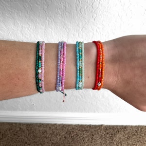 Op de afbeelding: Vijf kleurrijke kralenarmbanden om de pols van een persoon. De armbanden zijn gemaakt van kleine kralen in tinten groen, roze, lichtblauw en oranje. Elke armband heeft een enkele witte kraal in het midden.