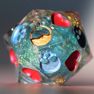 以下が含まれることがあります： 複数の面を持つ透明な樹脂製のサイコロで、ティール色のグリッター、金色のフレーク、さまざまなシンボルが埋め込まれています。シンボルには、赤いハート、青いスカル、黒い猫のシルエット、金色のポーションボトルが含まれます。このサイコロは、テーブルトップゲーム用に設計されています。