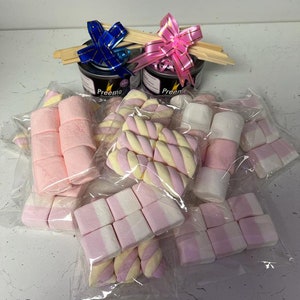 May include: A selection of packaged marshmallows in different shapes and colours, including pink and white, and pink and yellow swirl designs. Two small black containers with the word "Preemo" are topped with wooden skewers and blue and pink ribbons.