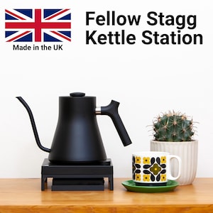 Op de afbeelding: Een zwarte Fellow Stagg waterkoker op een zwarte elektrische basis, een witte mok met een zwart en geel geometrisch patroon, een groene schotel en een cactus in een witte pot op een houten oppervlak. De tekst "Fellow Stagg Kettle Station" is zichtbaar boven de waterkoker. Een Union Jack-vlag met de tekst "Made in the UK" is zichtbaar in de linkerbovenhoek.