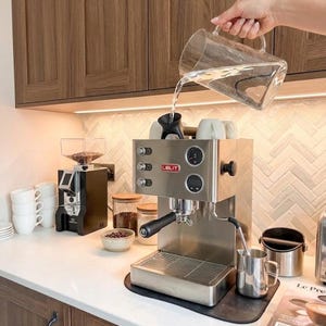 May include: A stainless steel Lelit espresso machine with a black portafilter, a drip tray, and a pressure gauge. Water is being poured from a clear glass pitcher into the top reservoir. A black coffee grinder and white ceramic mugs are nearby.