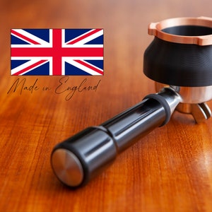 Op de afbeelding: Een zwarte en zilveren espresso portafilter met een koperkleurige bovenkant. De portafilter ligt op een houten oppervlak. Gemaakt in Engeland.