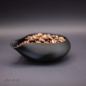 May include: A matte black ceramic bowl with a curved, teardrop-shaped pouring spout filled with roasted brown coffee beans. The bowl has a smooth, rounded exterior and a wide opening. The text "Made in the UK" is printed in the bottom left corner.