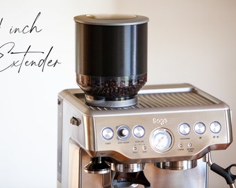May include: A silver espresso machine with a 4 inch extender on top. The extender is filled with coffee beans. The machine has a variety of buttons and a pressure gauge.