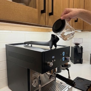 May include: A black espresso machine with a stainless steel portafilter and drip tray. A person is pouring water from a glass carafe into the machine. The machine has a pressure gauge and blue control knobs. The word "PROFIT" is visible on the machine.