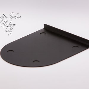 May include: Black metal coffee scale sliding tray with four holes. The tray is curved on one side and has a straight edge on the other side.  The text "Coffee Scale Sliding Tray" is written in cursive above the tray.