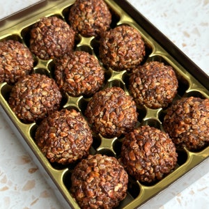 May include: A box of chocolate-covered treats with a crunchy texture. The treats are arranged in a grid pattern in a gold-coloured box with a brown lid.